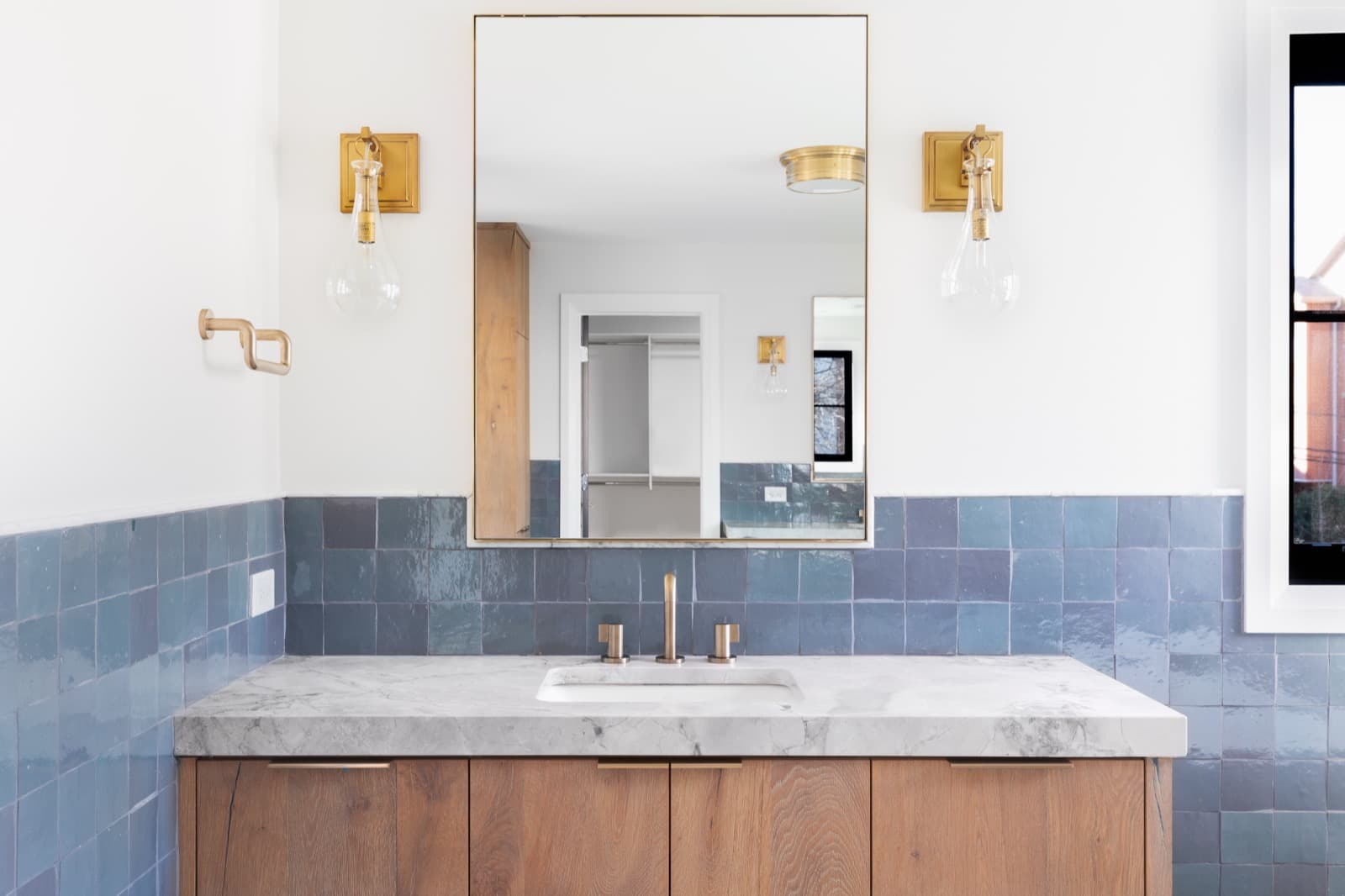 A bathroom vanity wall in blue zellige tile with a marble-topped oak vanity and brass sconces