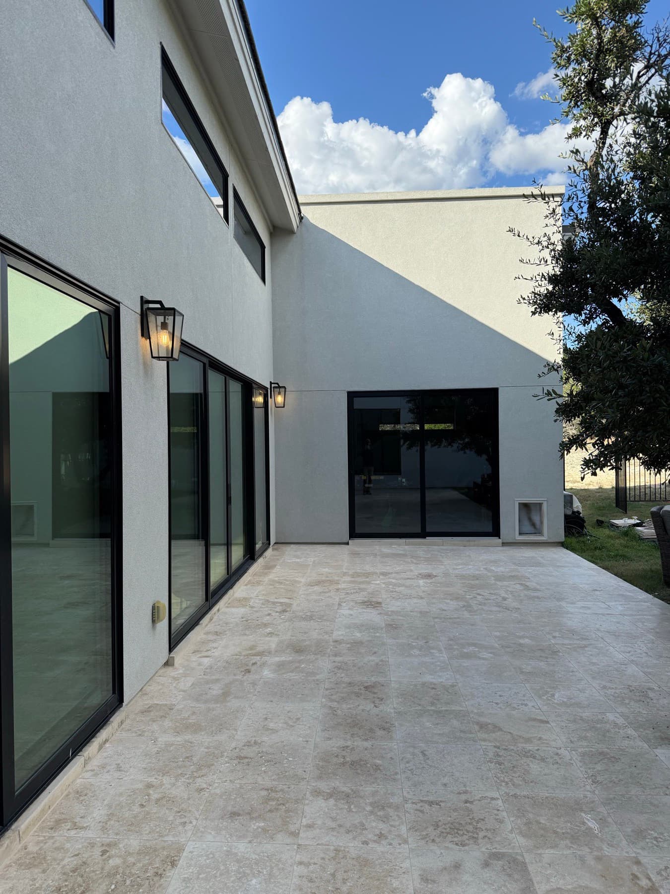 Travertine paving running the length of a white stucco house in Calabasas, past black-framed sliders, wall lanterns and an oak tree.