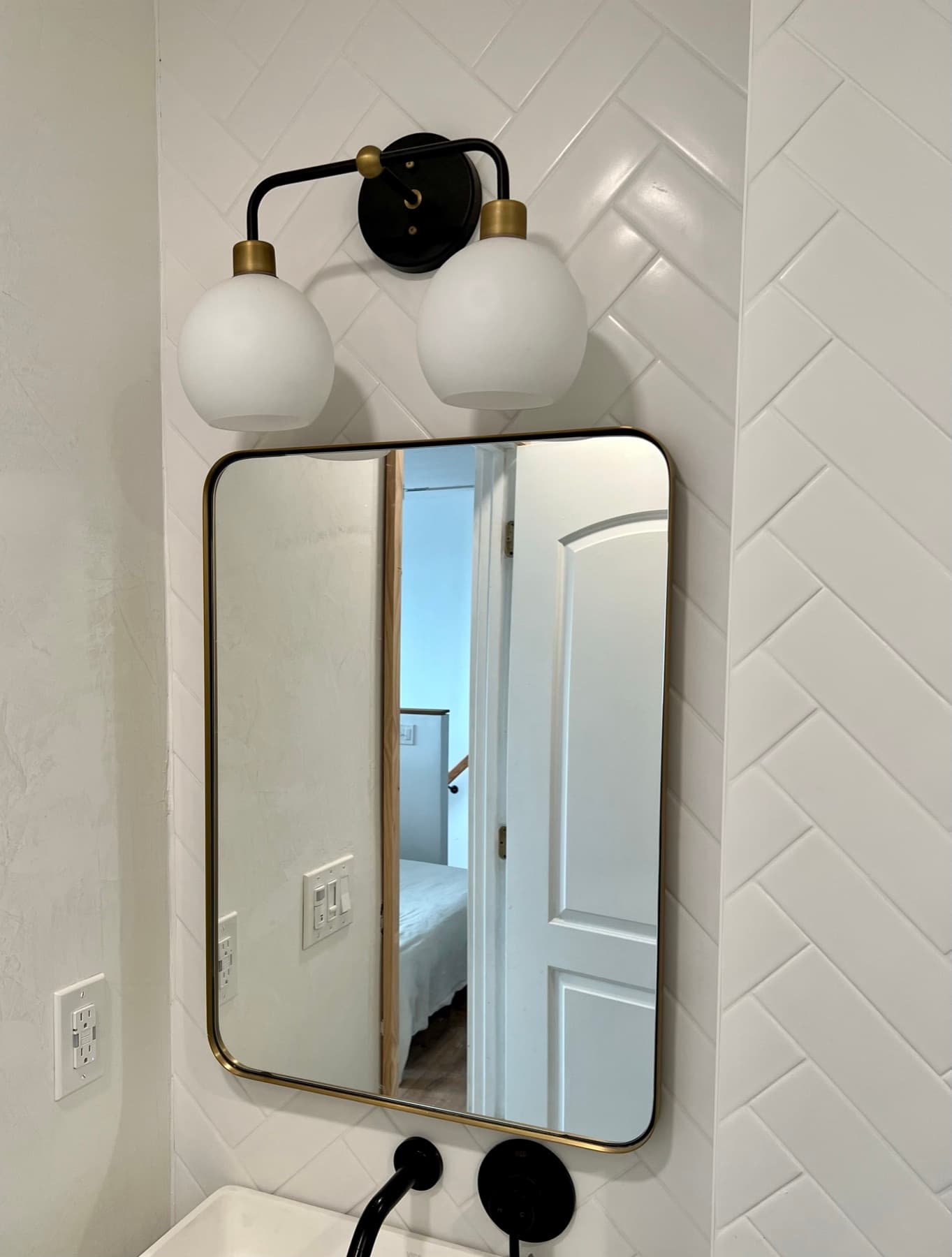 Brass-framed mirror and double globe sconce against the white herringbone wall