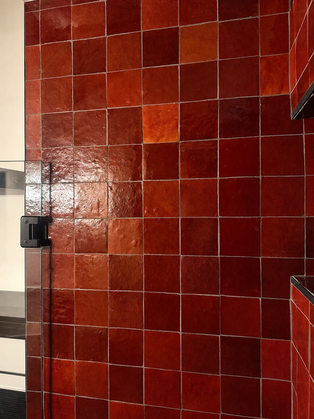 Close view of the rouge zellige wall beside the matte black glass door hinge, showing the shade variation in the handmade tile.