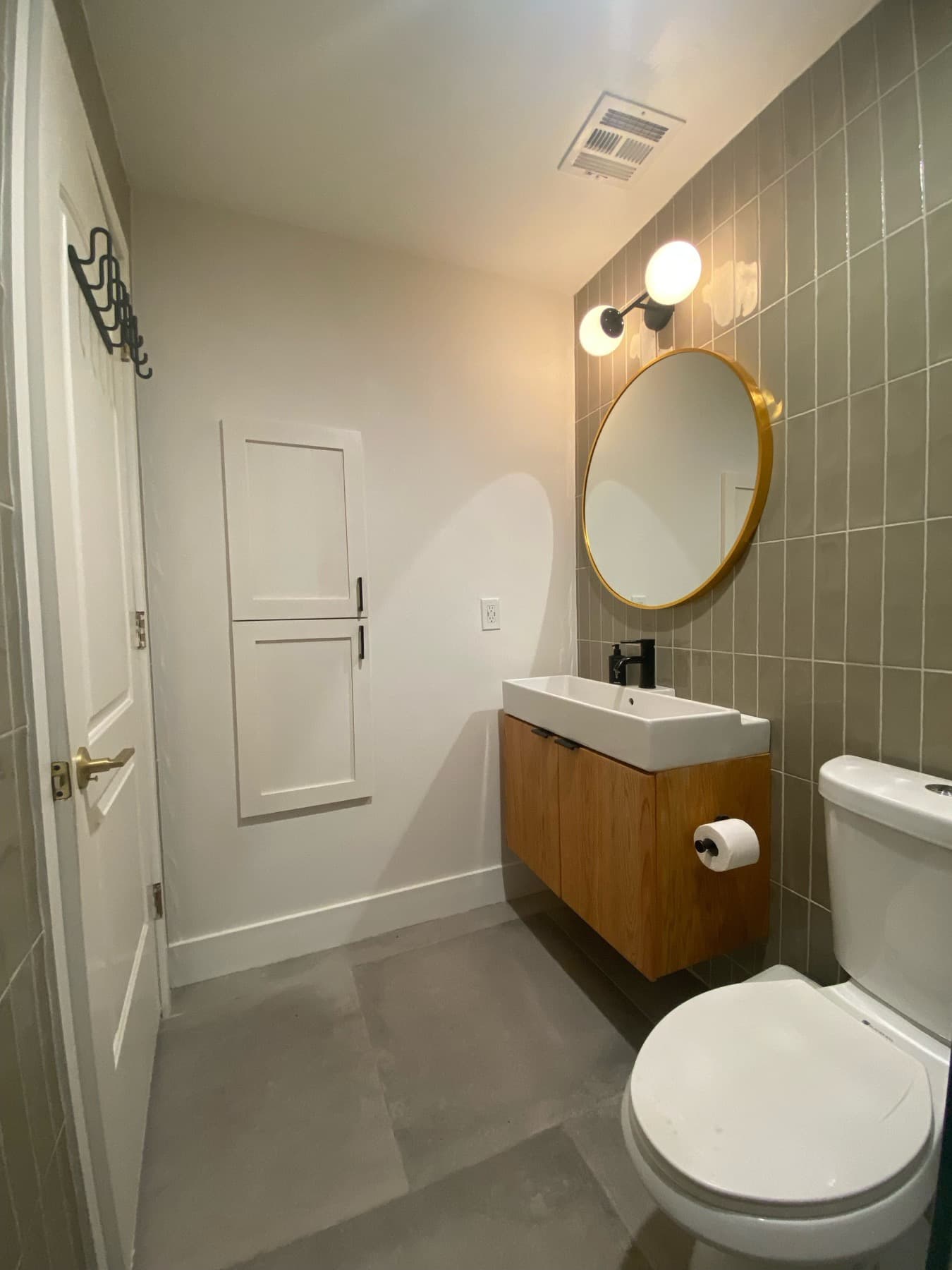 The floating oak vanity and round brass mirror, where the sage tile wall meets the painted wall by the door.